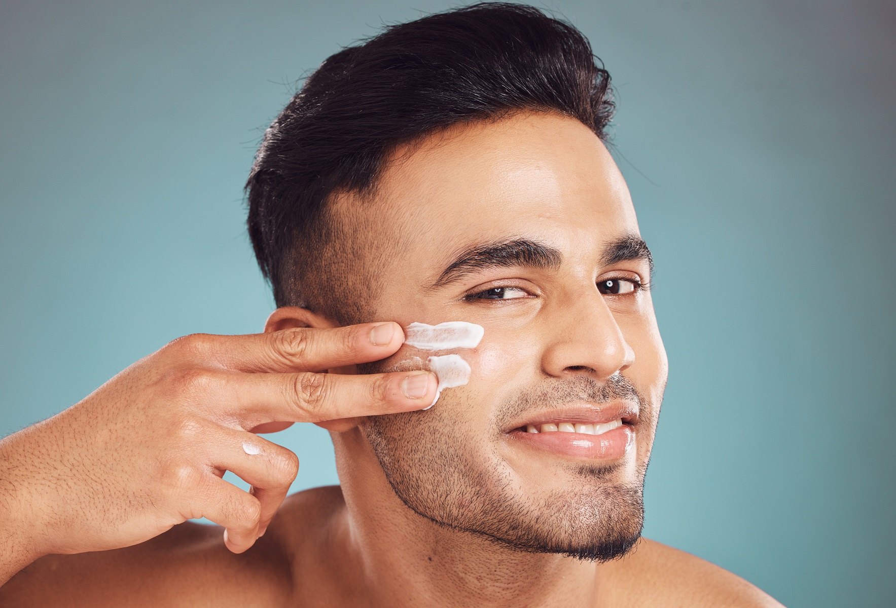 Portrait, beauty and cream with a man on a gray background in studio to apply antiaging face treatment. Facial, skincare and lotion with a young male person indoor for wellness or aesthetic self care. | Soleill Medical & Beauty Spa in Portland, OR